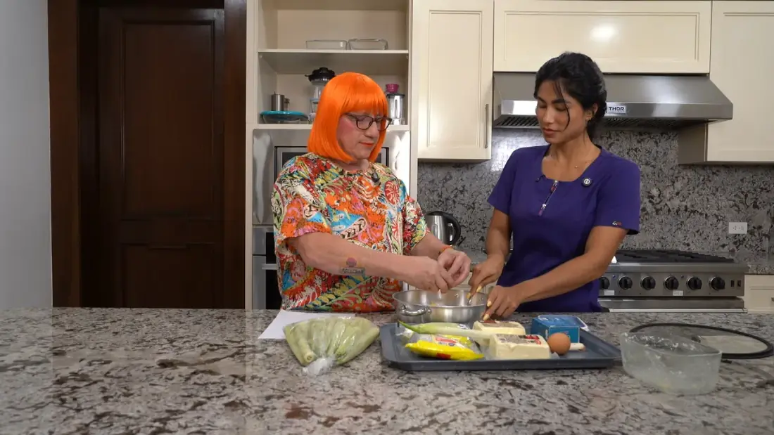 Un invitado con peluca naranja y la host partiendo huevos en una olla, con los ingredientes servidos en la mesada.